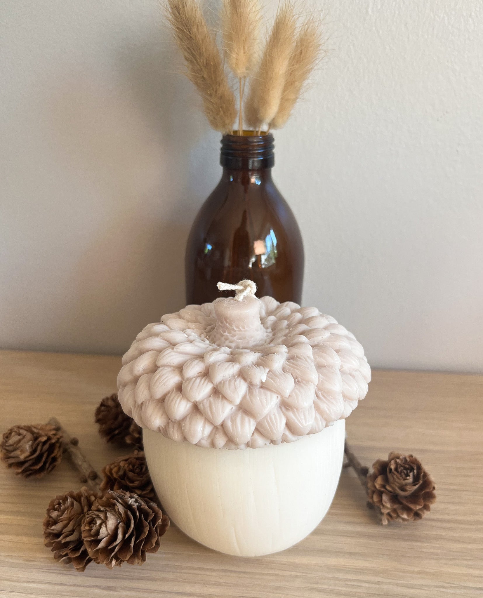 Beige ahorn sojalys med kogler og vase i baggrunden. Håndstøbt i sojavoks og inspireret af naturens farver.