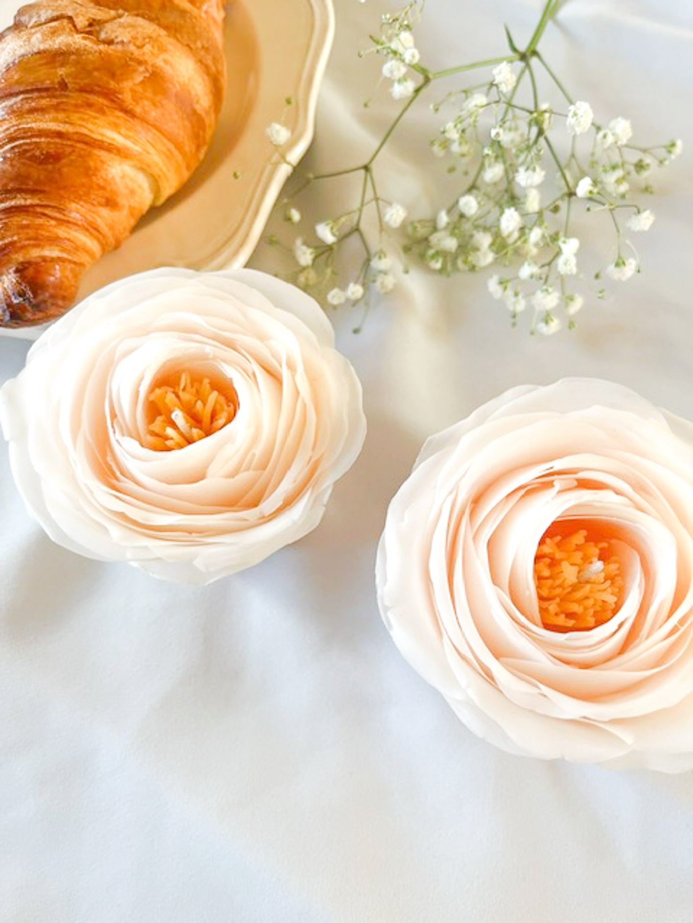 Et stemningsfulgt billede af to smukke rose blomsterlys i hvi med orange center og dele af en tallerken med en croissant og blomster.